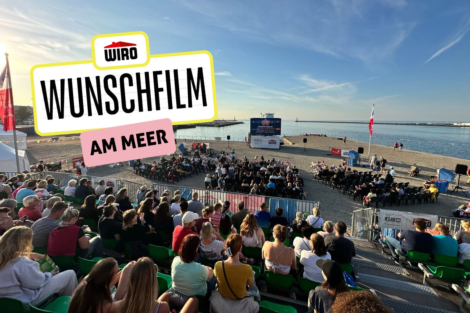 Blick von der Tribüne auf der Warnemünder Mittelmole auf die Ostsee - viele Menschen sitzen vor einer großen LED-Leinwand - auf dem Bild ist das Logo des WIRO-Wunschfilms