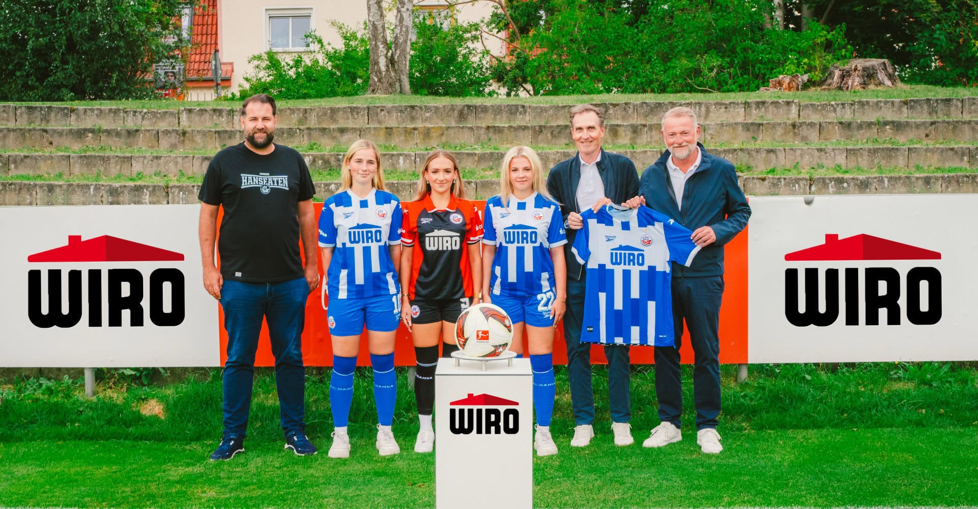 sechs Personen (drei Männer, drei Fußballspielerinnen) stehen im Rostocker Volksstadion vor einer Werbebande der WIRO, im Vordergrund eine Ball-Säule